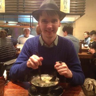 A man in a busy restaurant looking at the camera while stirring his food