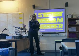 A woman standing in a classroom in front of a screen, looking at the camera