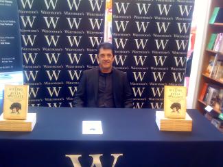 A man sitting at a table at a book launch, with books piled either side of him