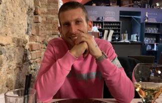 A man sitting at a restaurant table, with his elbows on the table and hands in front of his face, smiling