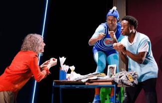 Actors performing in a theatre play, with a woman on the left speaking to a young man on the right. Another man is standing on a chair