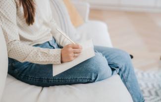 A woman sitting on a sofa writing in a diary. Her face is not in view