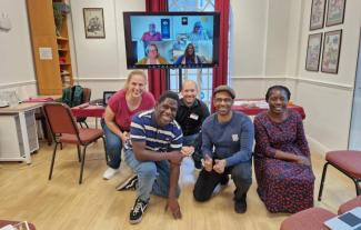 Five people crouching and smiling for the camera. Behind them is a video screen featuring people on a video chat