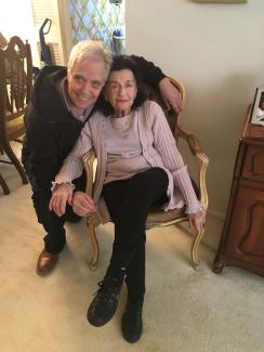 An elderly woman in a chair, with her son standing next to her, both smiling