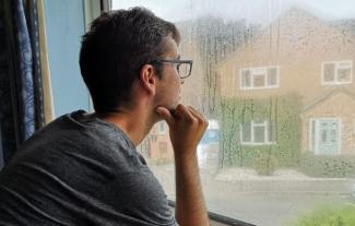 A young man looking out of a window on a rainy day, with his hand to his chin