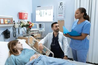 A female nurse standing beside a hospital bed, talking to a patient. A seated male doctor is listening.