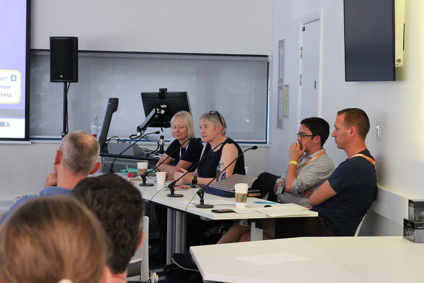 A woman at a desk addressing an audience, with several people sitting next to her