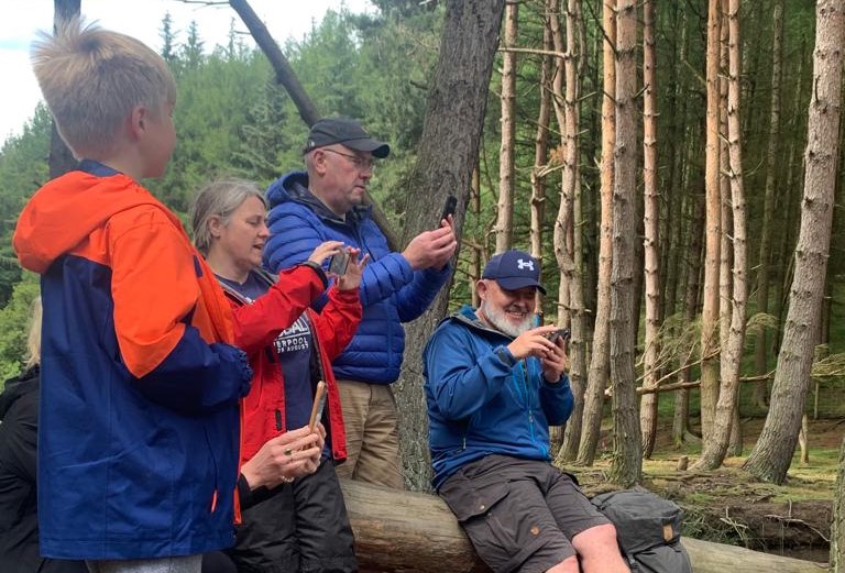 A child and four adults in a forest, some sitting, some standing, the adults are holding mobile phones